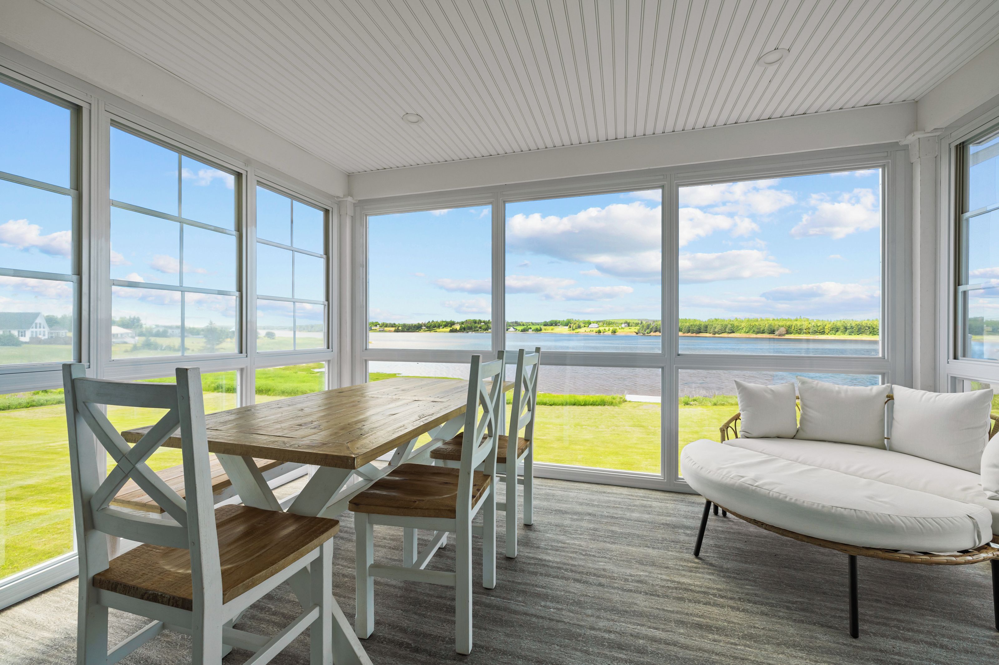 A room with a wooden table and chairs, a white sofa, and large windows offering a view of a body of water and greenery.