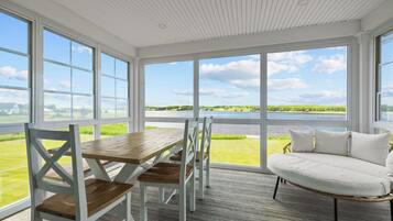 A room with a wooden table and chairs, a white sofa, and large windows offering a view of a body of water and greenery.