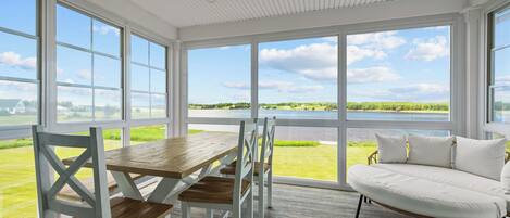 A room with a wooden table and chairs, a white sofa, and large windows offering a view of a body of water and greenery.
