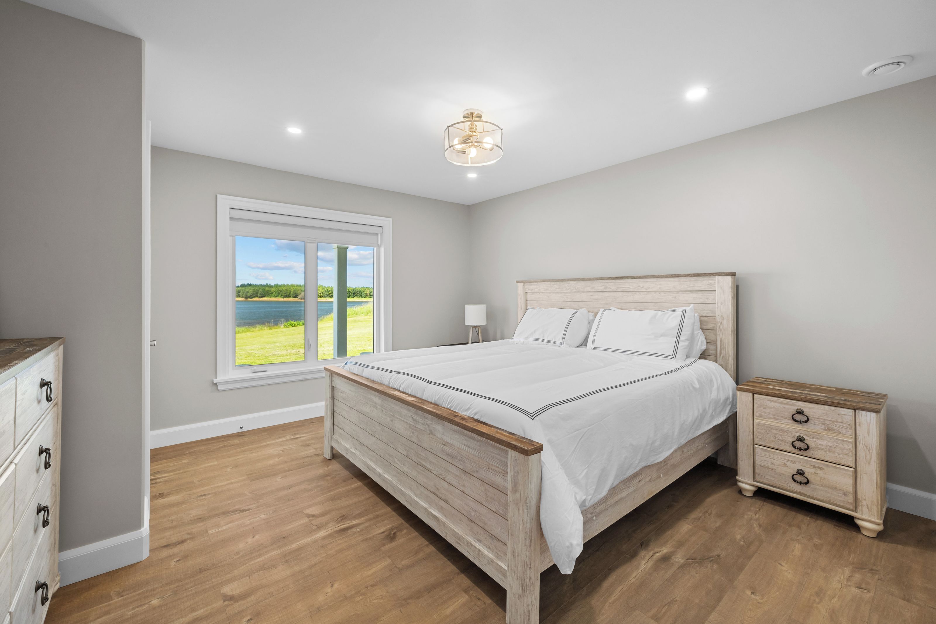 A bedroom with a wooden bed, a nightstand, a dresser, and a large window with a view of greenery.