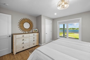 A bedroom with a large bed, a dresser with drawers, a mirror, and a view of a lake through a window.