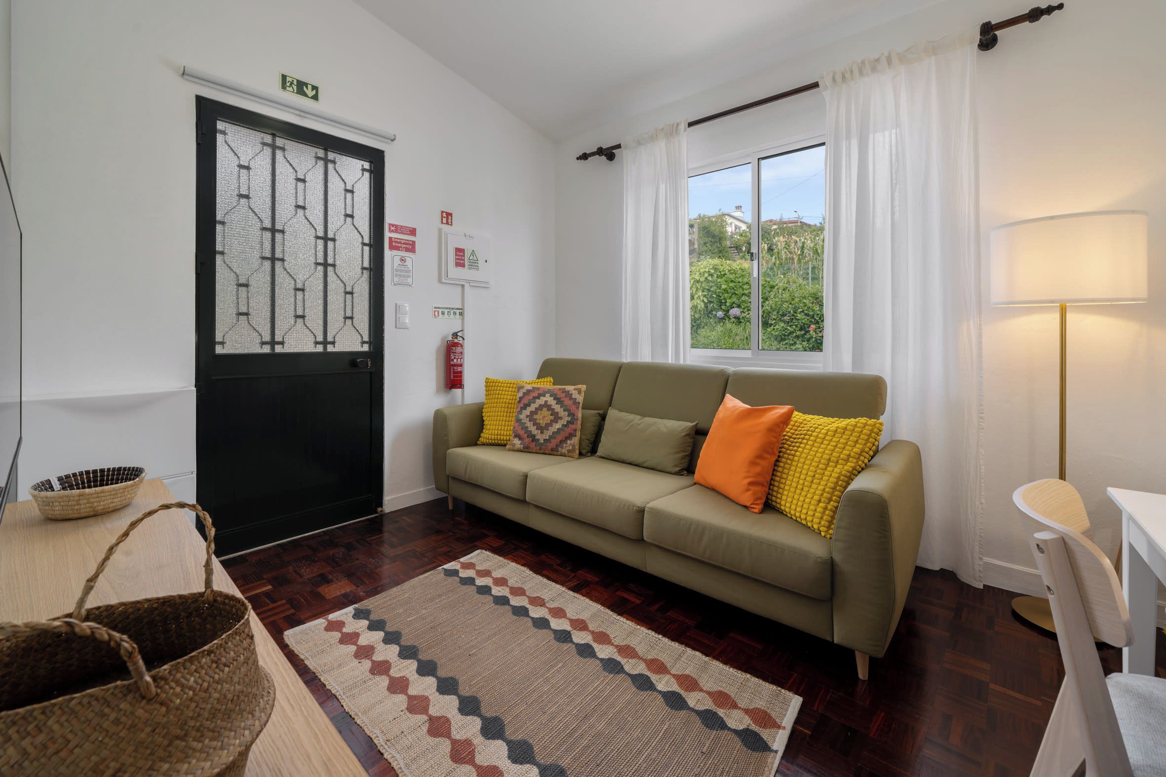A cozy living room with a sofa, a patterned rug, a window with curtains, and a door with emergency exit signs.