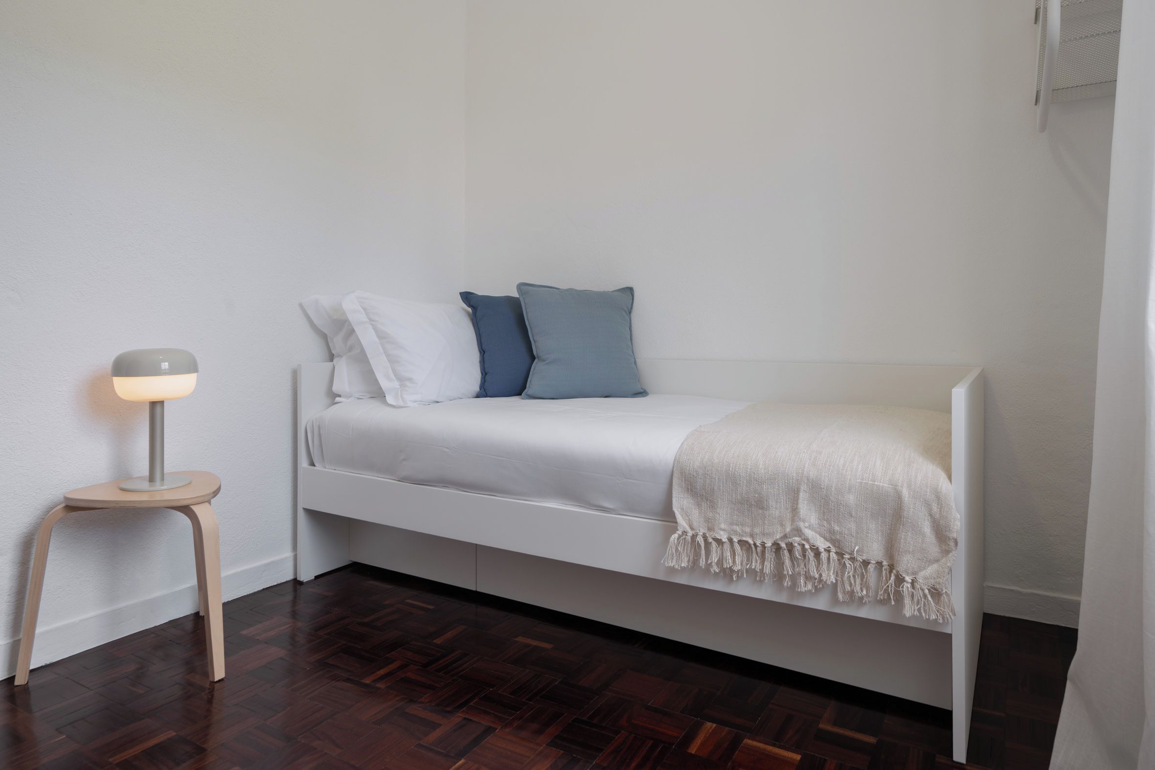 A small, single-bed room with a white bed, a bedside lamp, and a wooden floor.