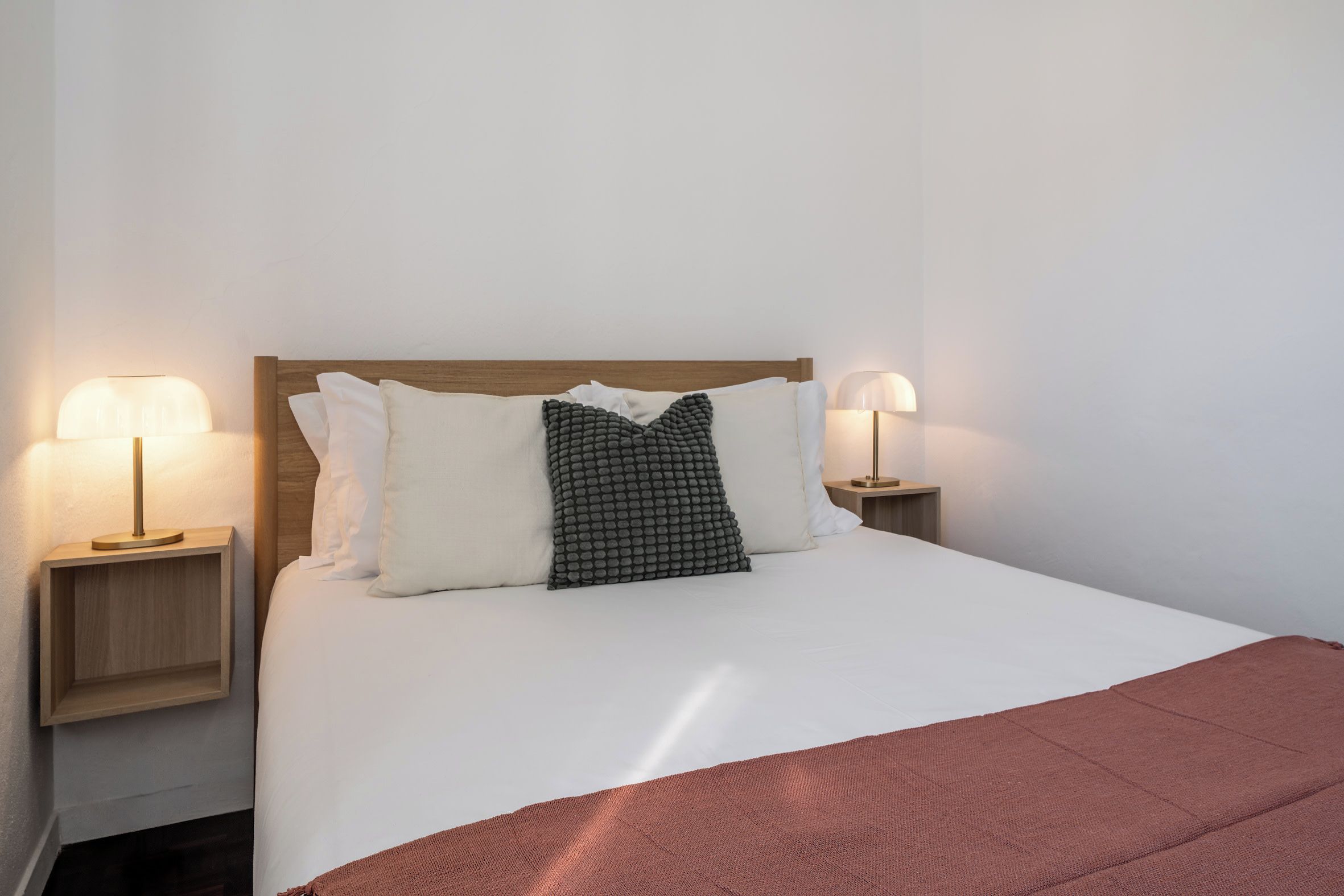 A neatly made bed with white bedding and a brown throw blanket, flanked by two bedside tables with lamps, against a plain wall.