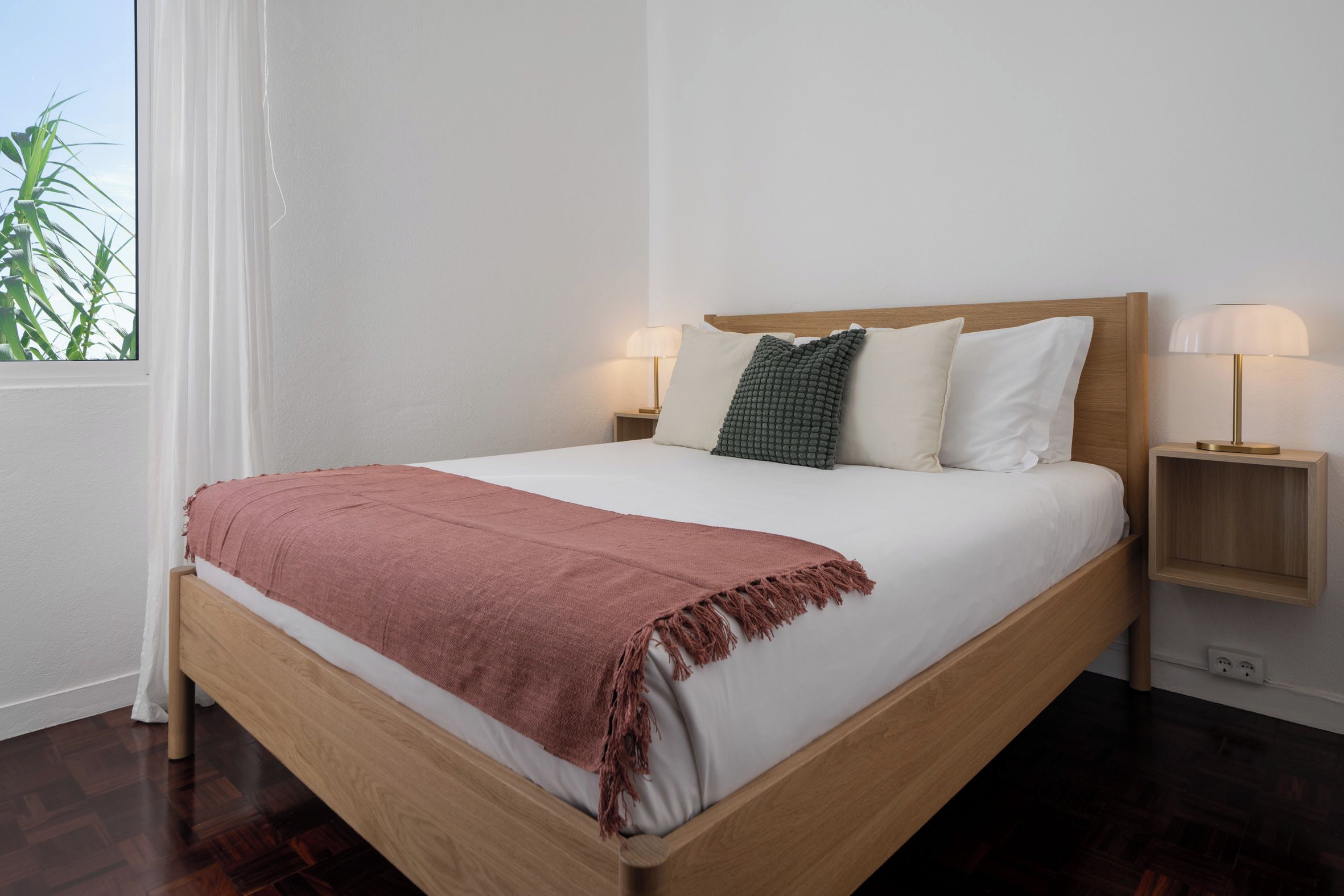 Una cama con estructura de madera, ropa de cama blanca, un plaid rosa y dos lámparas de noche.