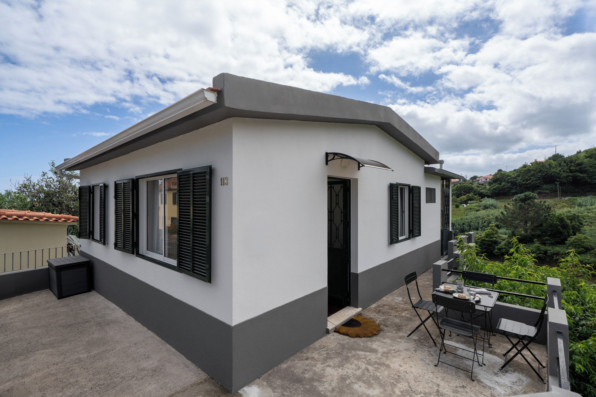 Een modern huis met twee verdiepingen, een balkon, een klein terras en uitzicht op een groene heuvel.