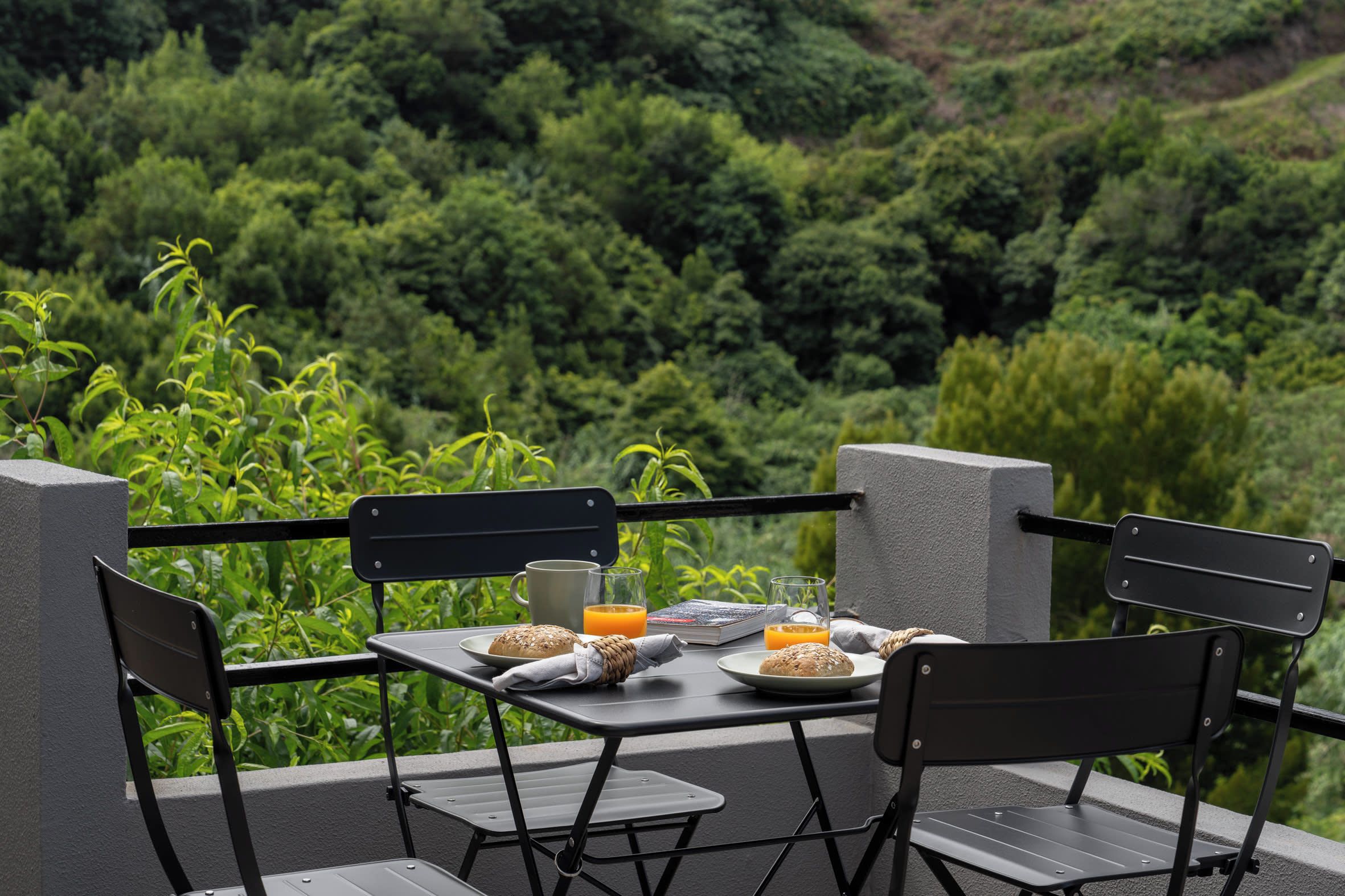 A table set outdoors with chairs, a book, a glass of orange juice, a plate with bread, and a cup.