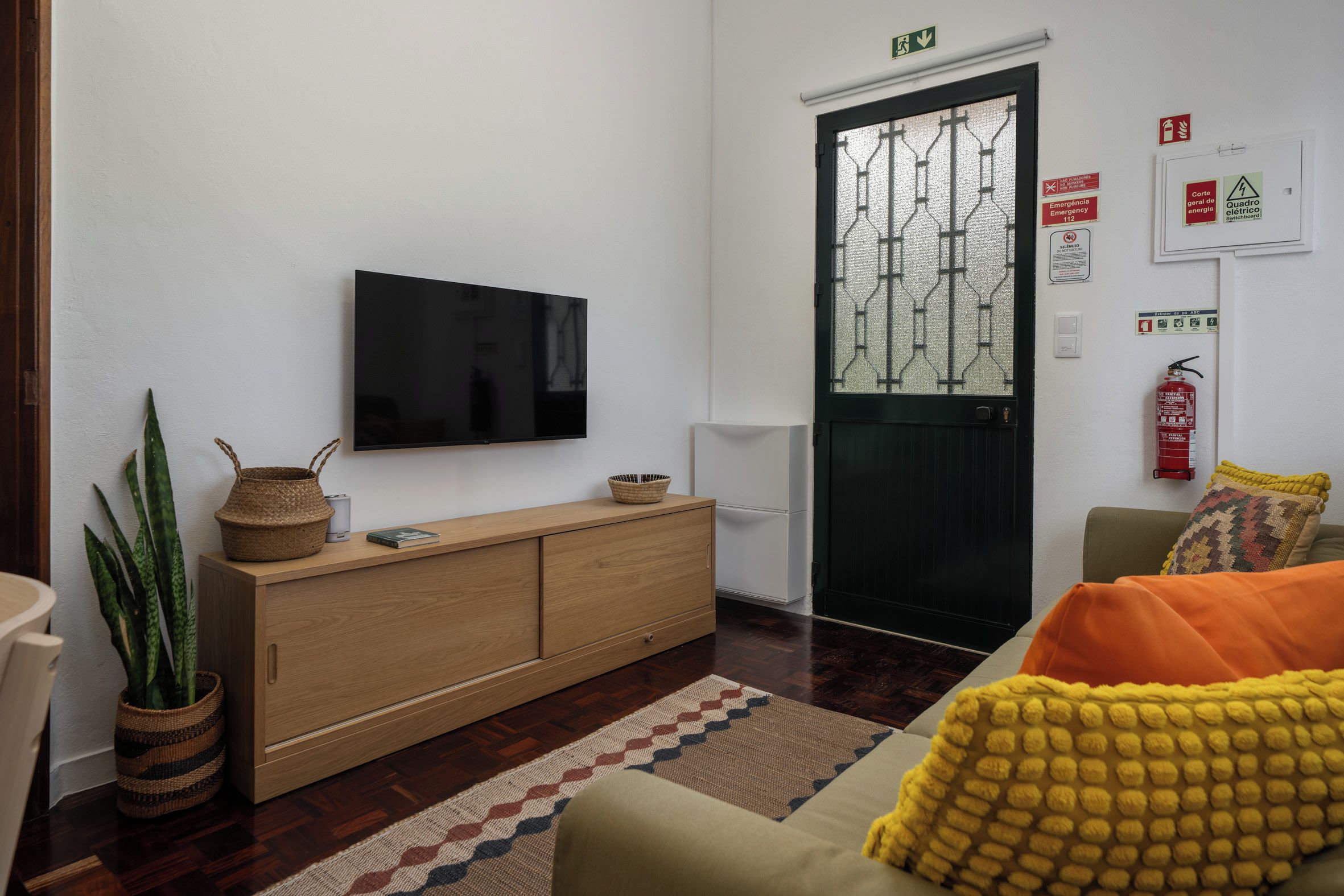 Un salon moderne avec un téléviseur à écran plat fixé au mur, un meuble en bois avec un panier tressé, un tapis à motifs et un canapé agrémenté d’un coussin jaune.