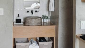 Ein Badezimmer mit einem runden Waschbecken, einem Spiegel und einem Handtuchhalter. Auf der Ablagefläche stehen Holzaufbewahrungsboxen und ein kleiner Topfpflanze.