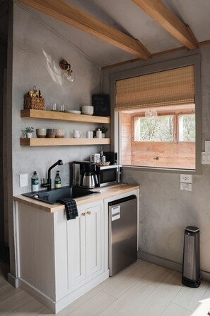 A kitchen with wooden shelves, a coffee maker, a microwave, and a window with blinds. - The A-Frame · Hot Tub, Fire Pit, 30 min to Waco (Valley Mills)