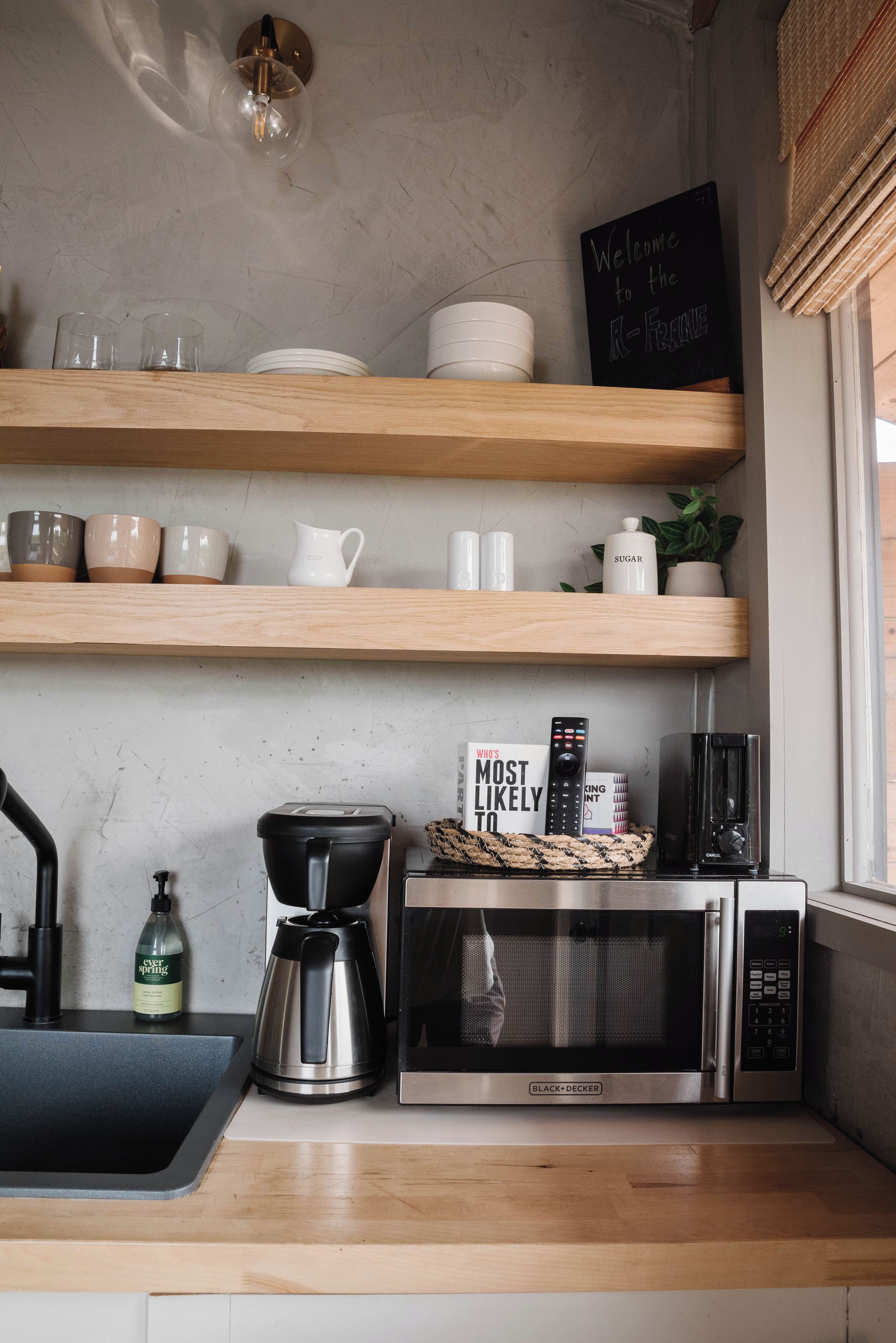 A kitchen with wooden shelves holding dishes, a coffee maker, a microwave, and a sign that says 'Welcome to the A-Family.'