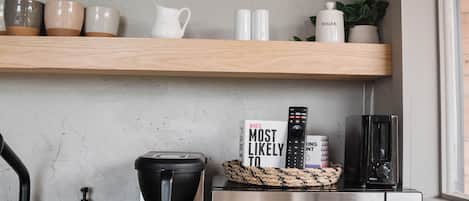 A kitchen with wooden shelves holding dishes, a coffee maker, a microwave, and a sign that says 'Welcome to the A-Family.'
