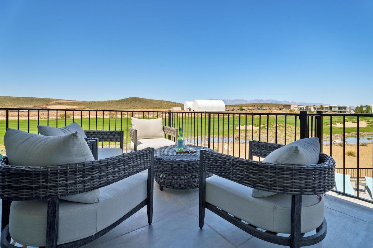 A balcony with wicker furniture, a table, and a view of a green field and mountains.