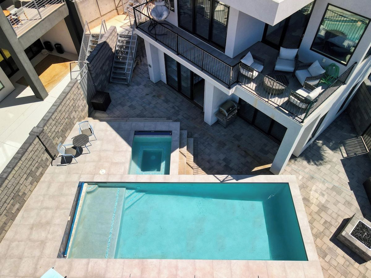 A rooftop pool area with a large pool and a smaller pool, surrounded by outdoor seating and a staircase leading to a balcony with a seating area.