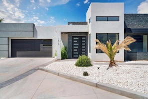 Una casa moderna de dos pisos con garaje, una puerta principal negra y una palmera pequeña en el jardín delantero.