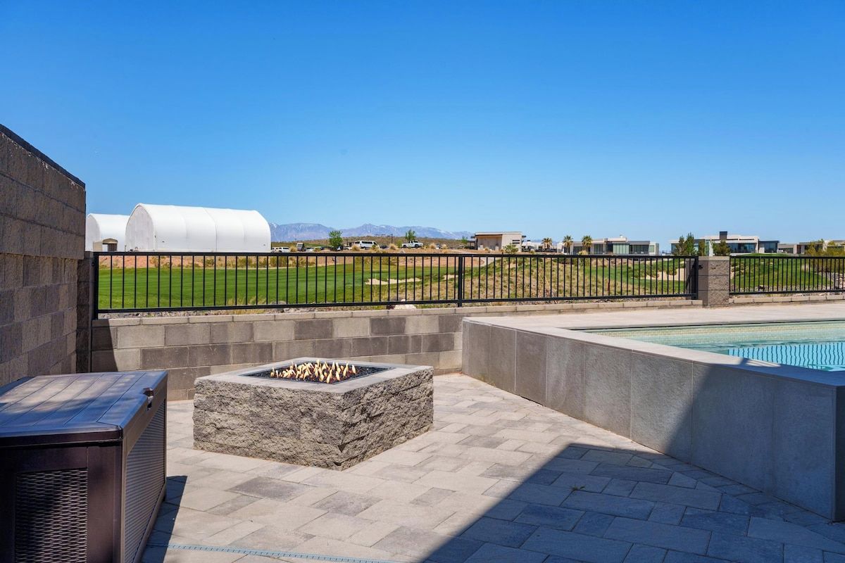 An outdoor patio area with a stone fire pit, a pool, and a view of a sports field.