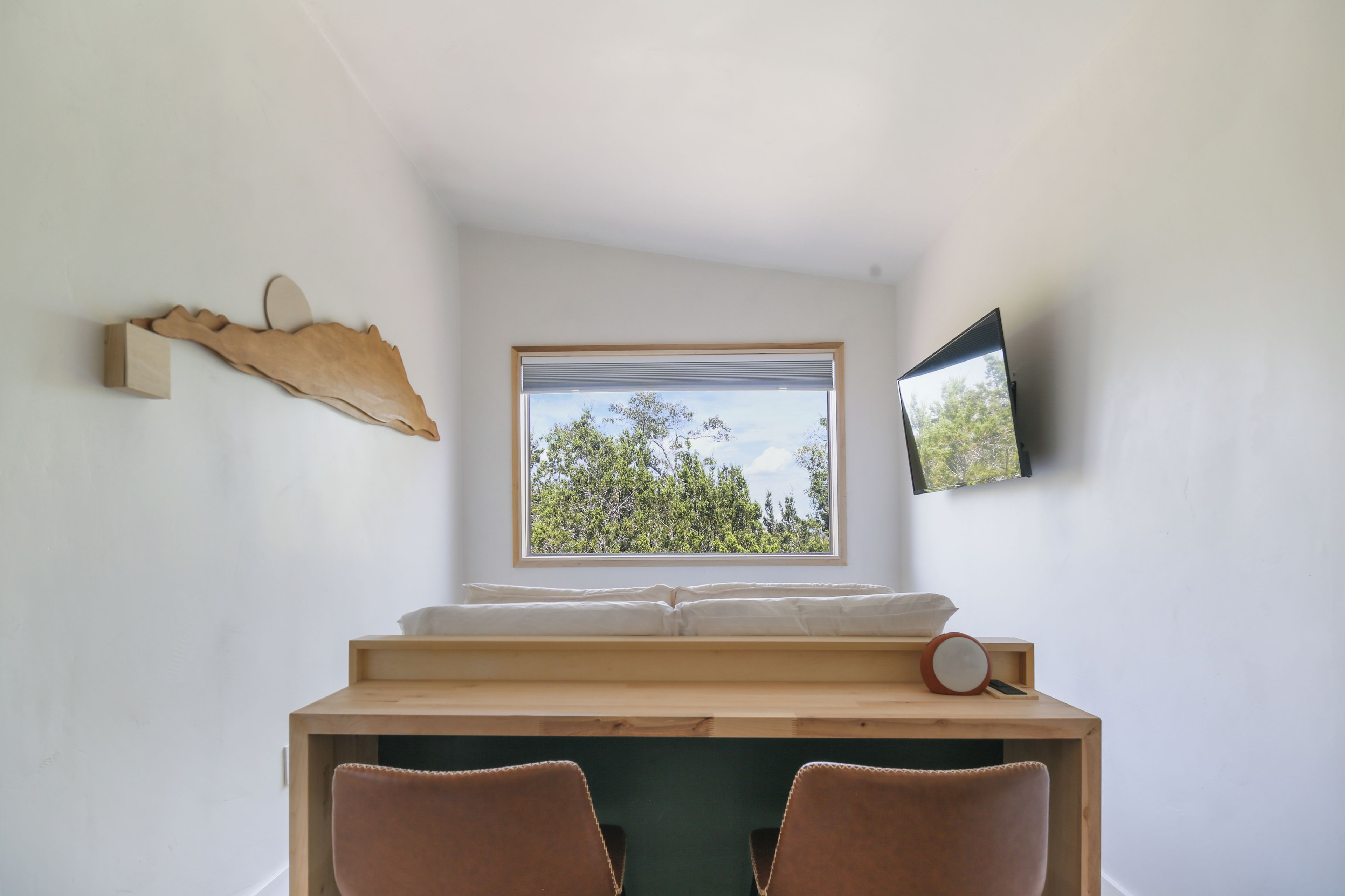 Une chambre avec un lit, un bureau, une chaise, une fenêtre donnant sur des arbres et un téléviseur fixé au mur.