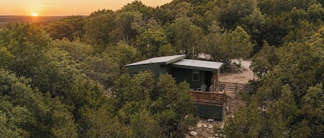 A small cabin surrounded by dense forest, with a clear view of the sunset in the background.