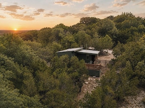 A small cabin surrounded by dense forest, with a clear view of the sunset in the background.