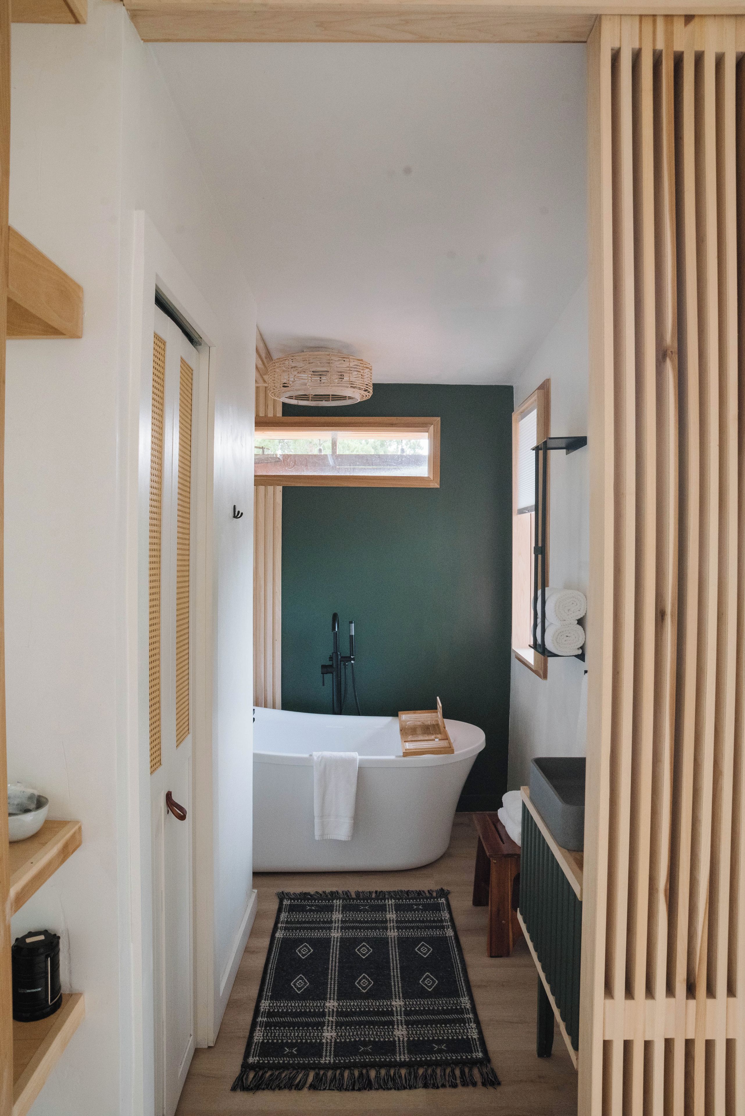 A narrow hallway leading to a bathroom with a freestanding bathtub, a window, and a wooden door.