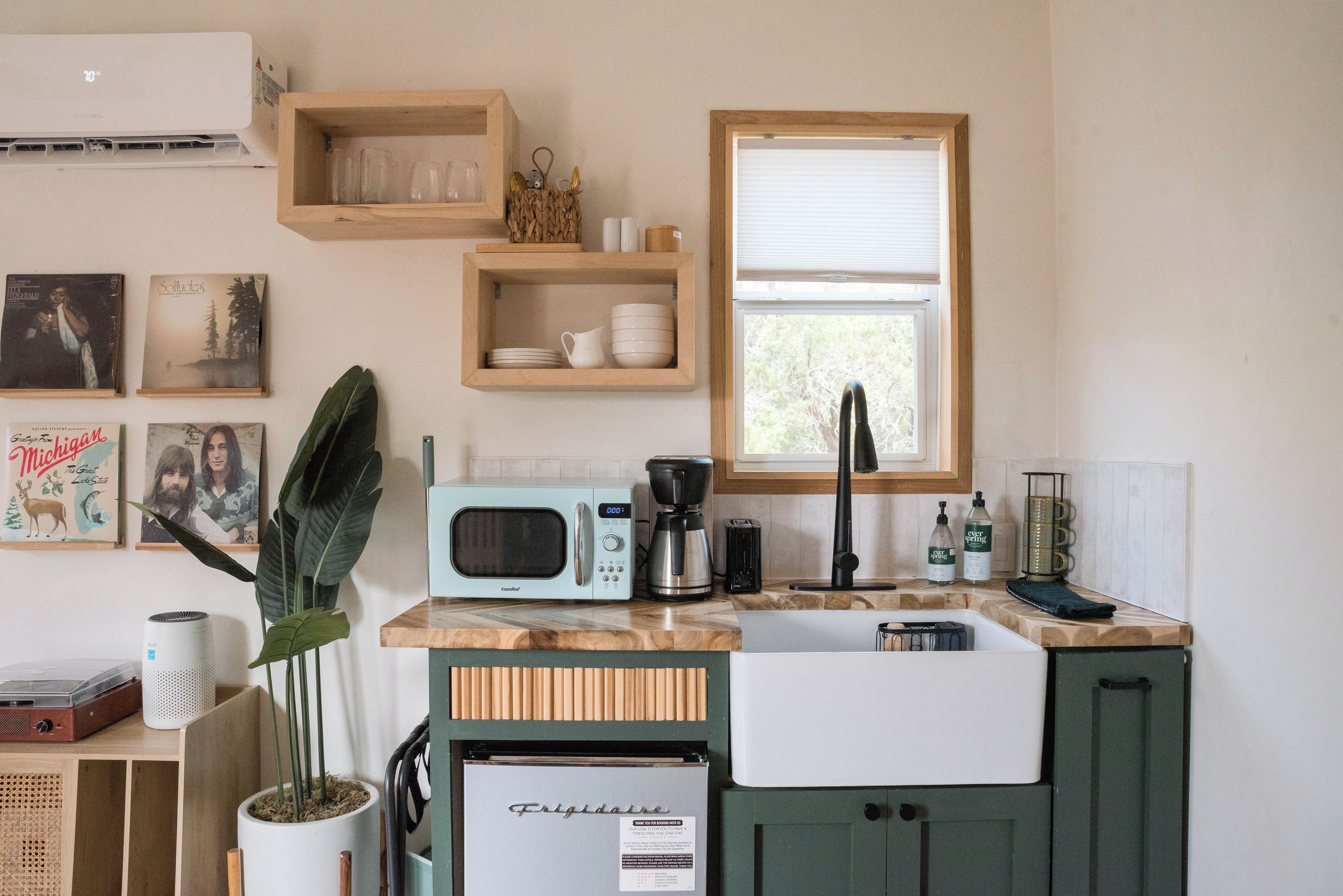 A compact kitchen with a microwave, coffee maker, and a refrigerator. There are shelves with dishes and a window providing natural light.