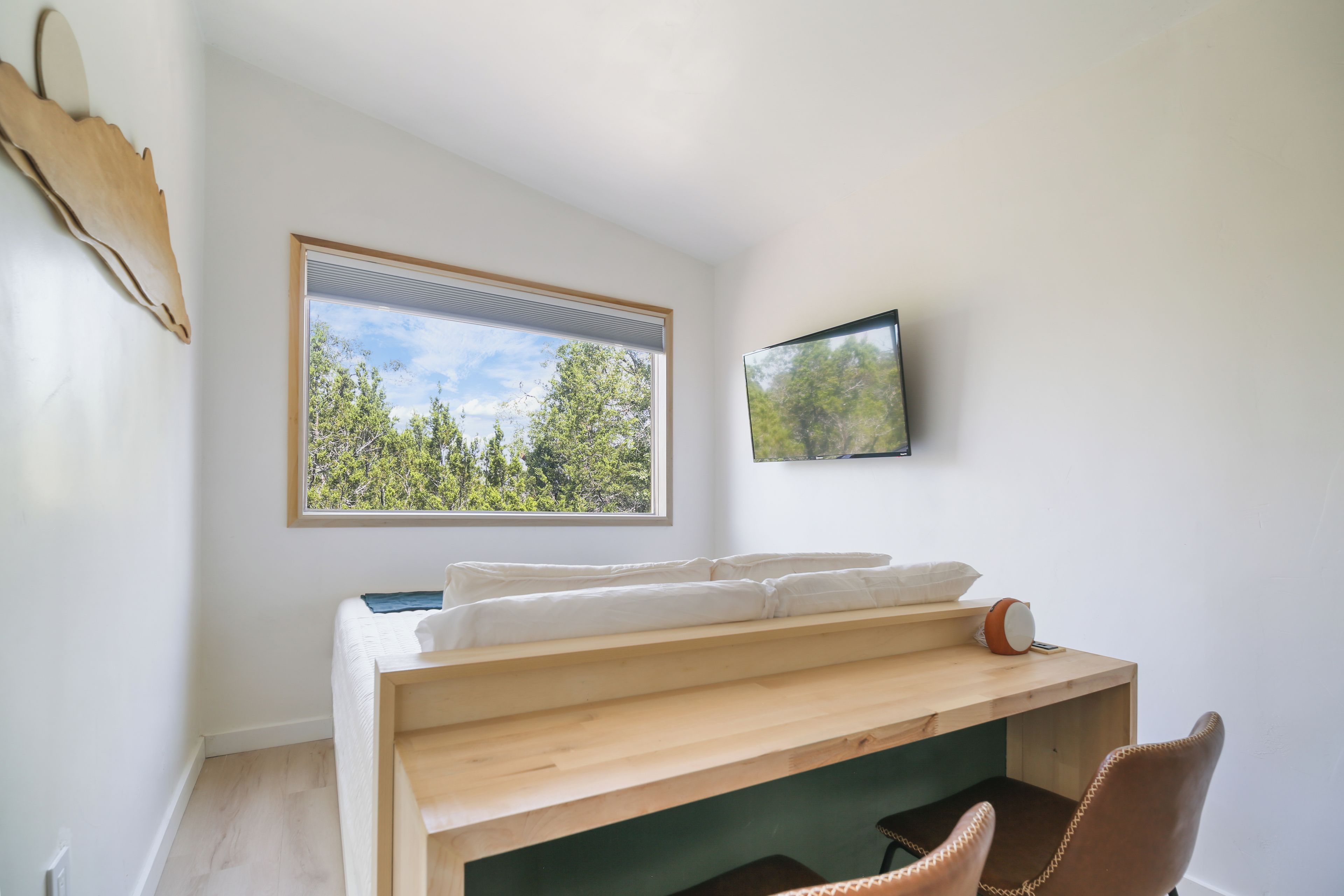 A room with a bed, a desk, a chair, a window with a view of trees, and a mounted television.
