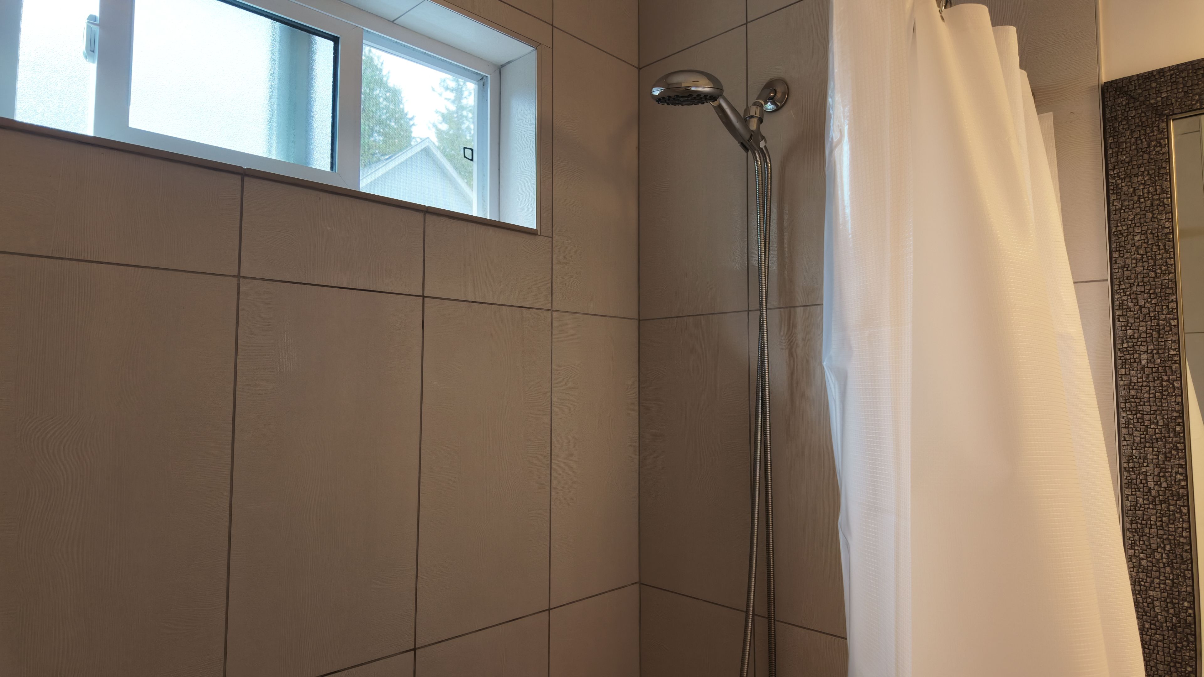 Una ducha con cabezal de ducha de mano, una ventana encima y una cortina medio bajada.
