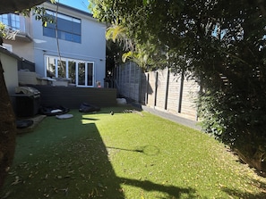 A backyard with artificial grass, a wooden fence, and a house with large windows. - 4-bedroom house in amazing Dover Heights with cool AC (Dover Heights)