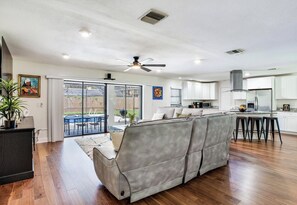 A spacious living room with a grey sofa, wooden flooring, a ceiling fan, and a sliding glass door leading to an outdoor patio. - Private Pool Retreat, Spacious 3BR + Large Yard (Brandon)
