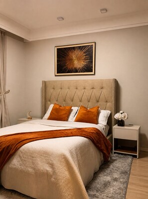A bedroom with a tufted headboard, a large bed with a beige and orange bedspread, a nightstand with a vase of flowers, and a wall-mounted abstract painting. - 2BR Near JKIA with Pool, Gym, Security, amenities & Free Parking (NAIROBI)