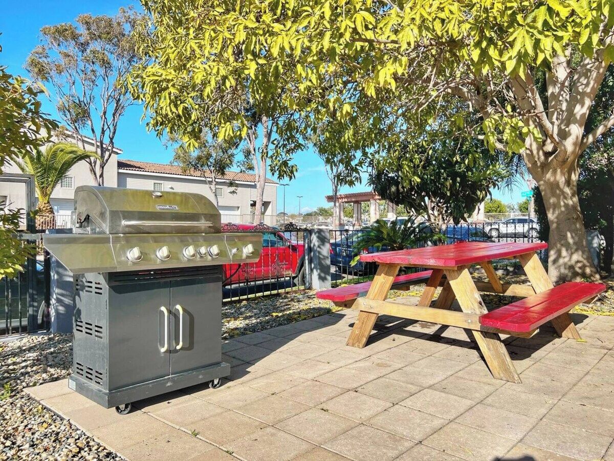 An outdoor barbecue grill and picnic table in a sunny area with buildings and parked cars in the background.
