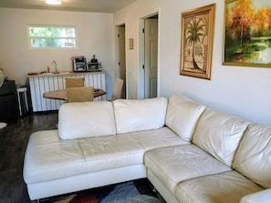 En stue med en hvid lædersofa, et spiseområde med bord og stole, og et køkken med hårde hvidevarer.