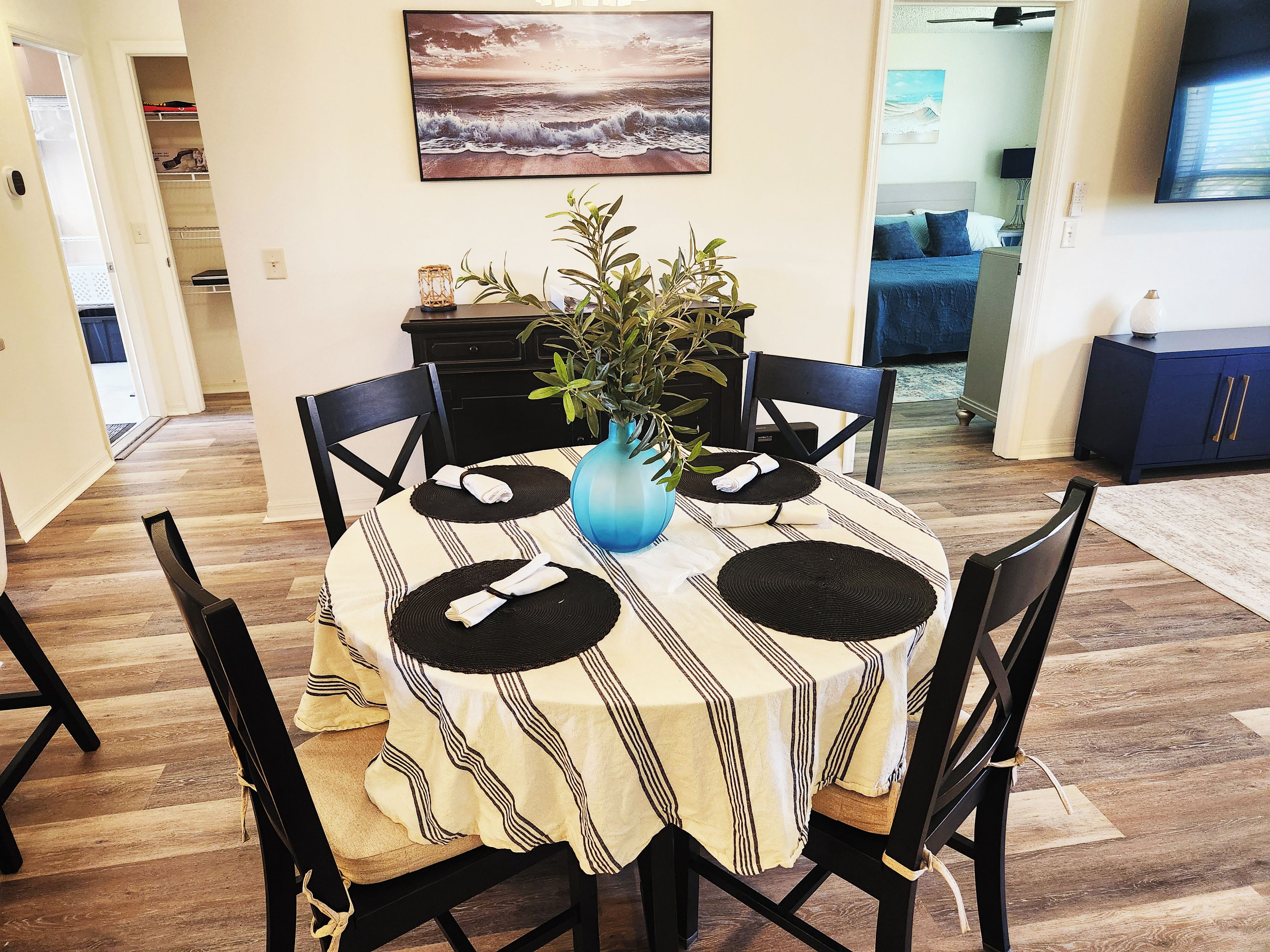 A dining area with a round table, black chairs, a striped tablecloth, and a blue vase with a plant.