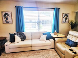 A living room with a beige sofa, a small side table, and a window with blue curtains. There are two framed seahorse artworks on the walls. - Coastal Breeze Getaway 2BR/2BA BBQ + Fire Table + Lanai + Garage (The Villages)