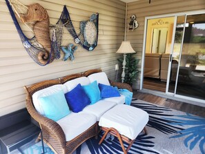 A wicker sofa with white and blue cushions, a potted plant, and a lamp on a porch with a glass door leading to a seating area. - Coastal Breeze Getaway 2BR/2BA BBQ + Fire Table + Lanai + Garage (The Villages)