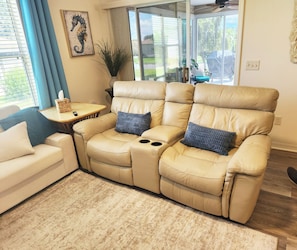 A beige leather sofa with a reclining feature, a small wooden side table, and a blue cushion. - Coastal Breeze Getaway 2BR/2BA BBQ + Fire Table + Lanai + Garage (The Villages)