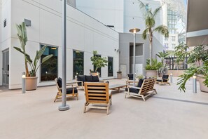 Un espace extérieur aménagé avec des chaises et des tables en bois, entouré de plantes en pot et bordé par la façade d’un bâtiment moderne.