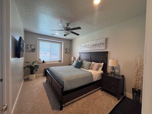 Une chambre à coucher comprenant un lit, des tables de chevet, un ventilateur de plafond, une fenêtre avec des stores et des œuvres d’art murales.