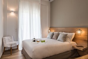 Una cama bien hecha con una bandeja de manzanas y una taza, una mesita de noche con una lámpara y una silla en una habitación con cabecera de madera y cortina translúcida.