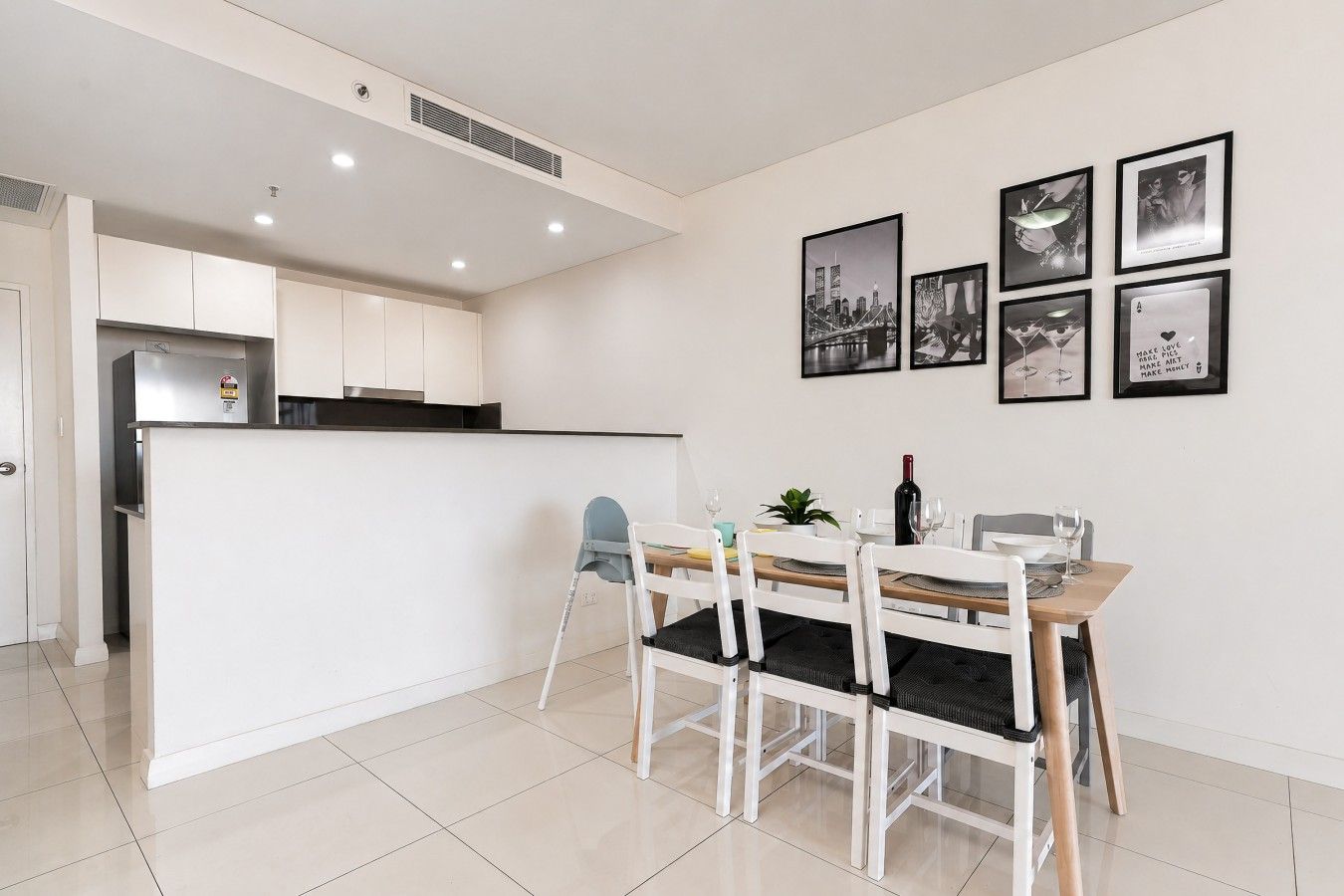 Une cuisine moderne avec des meubles blancs, un coin repas doté d’une table et de chaises en bois, et un mur orné de tableaux encadrés.