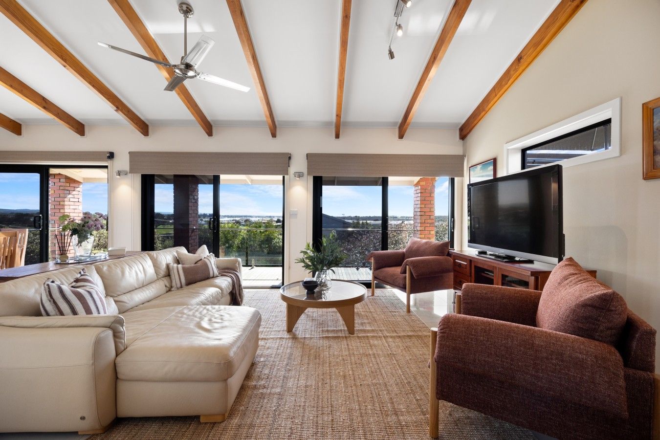 A spacious living room with a large sofa, a smaller sofa, a coffee table, and a television. The room has wooden beams on the ceiling and large windows offering a view of the outdoors.
