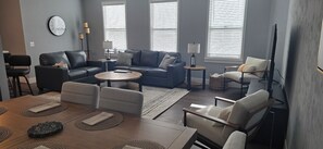 Una sala de estar moderna con sofá, mesa de centro y sillas, una mesa de comedor con individuales, y un ventanal con persianas.