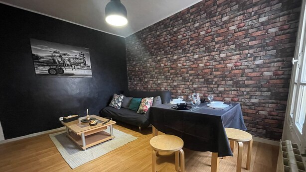 A room with a brick wall, a black sofa, a dining table set for two, and a wooden floor. - Cozy, near Paris (Villeneuve-Saint-Georges)