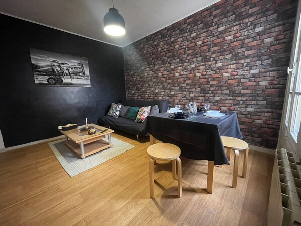 Une pièce avec un mur en briques, un canapé noir, une table à manger dressée pour deux personnes et un plancher en bois.