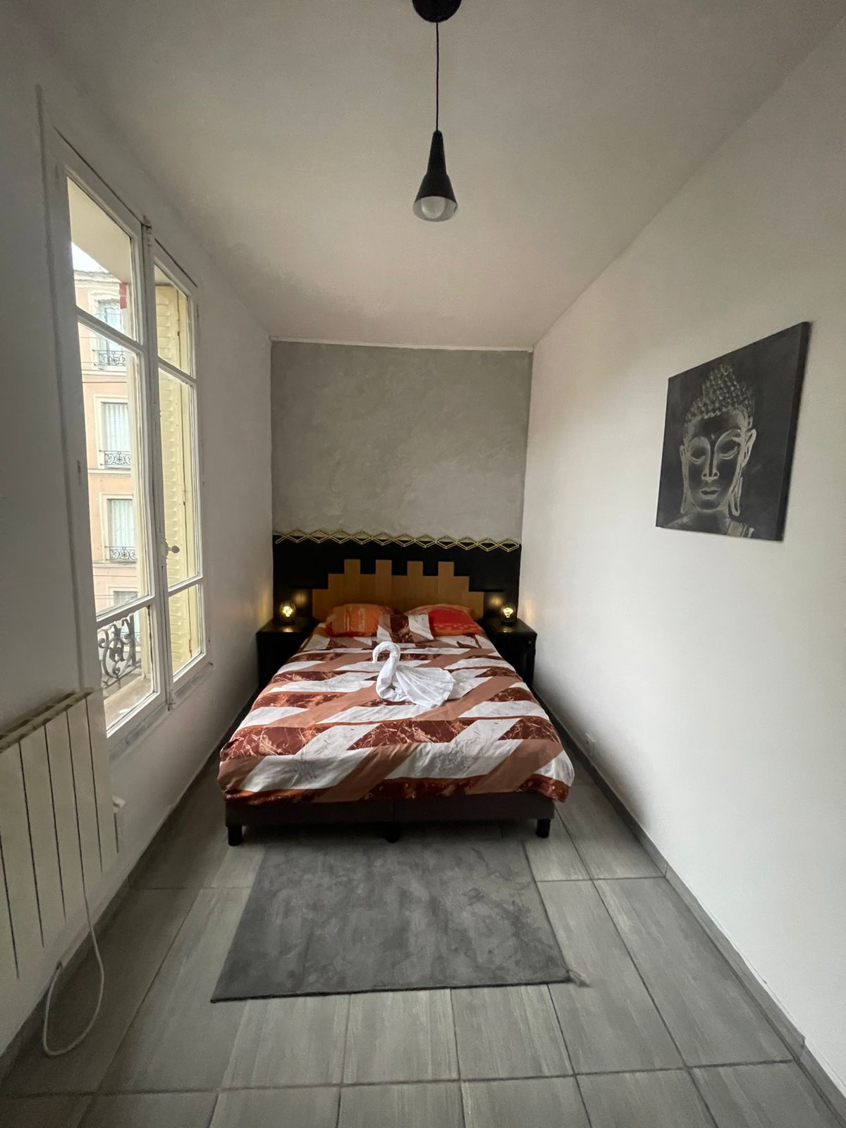 Een smalle hotelkamer met een bed, hoofdeinde, een kunstwerk aan de muur en een raam.