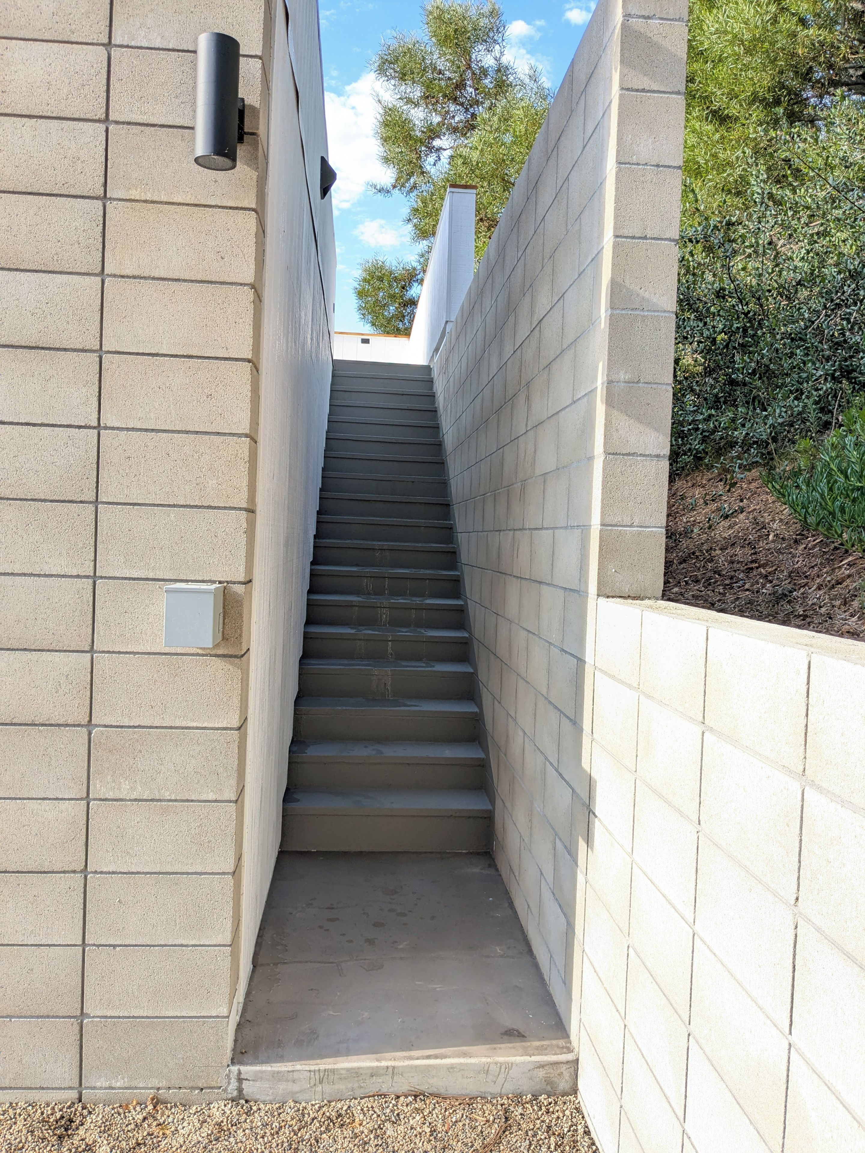 A concrete staircase between two walls with a white electrical box on the left and a small window at the top.