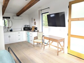 A compact kitchen with white cabinets, a microwave, and a coffee machine. There's a dining area with a wooden table and a wicker chair.