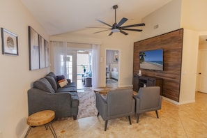 A living room with a sofa, chairs, a coffee table, a TV mounted on a wooden wall, and a ceiling fan. - Peaceful 3BR 2BTH Villa | Private Pool | Game Room | Champions Gate Area (Davenport)