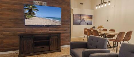 Una sala con un televisor de pantalla plana montado en una pared de madera, un comedor con una mesa y sillas de madera, y una lámpara colgante.