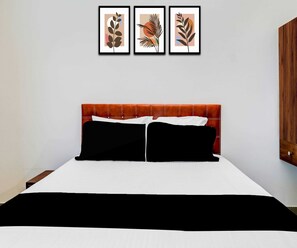 Ein Bett mit weiß-schwarzer Bettdecke, zwei schwarzen Kissen und einem braunen Kopfteil. Darüber hängen drei gerahmte Kunstwerke mit abstrakten Pflanzenmotiven.